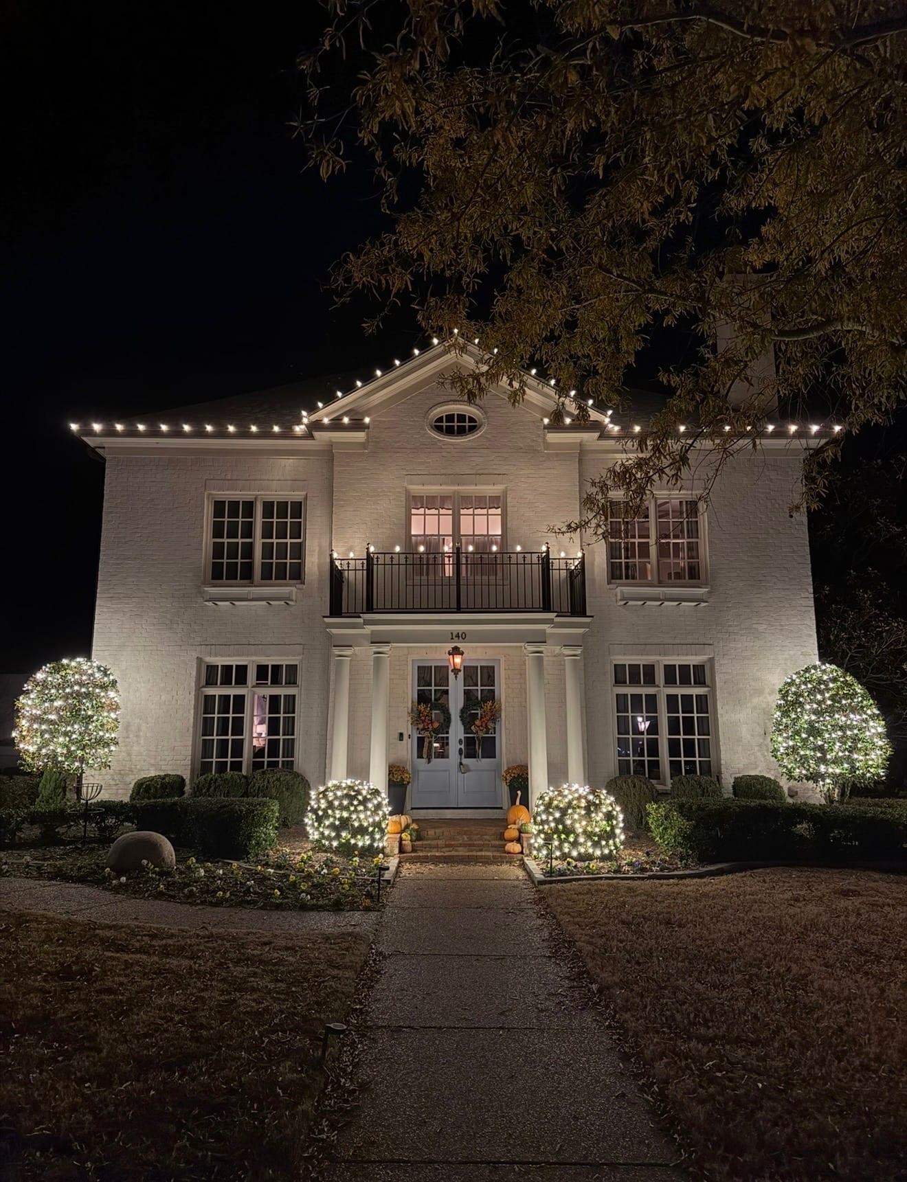 White-brick colonial at night with a warm white C9 roofline, two glowing lit spheres flanking the front walk, and net-lit topiary trees on either side of the entrance