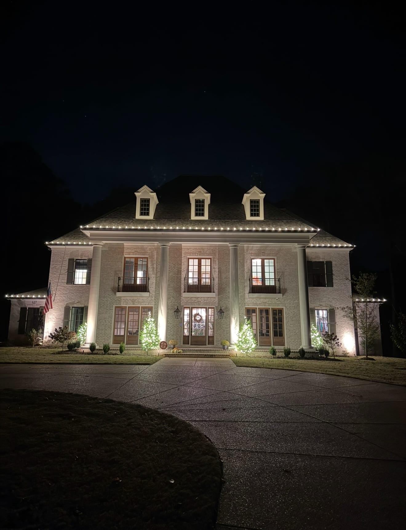 Grand two-story estate at night with warm white C9 bulbs along the roofline, an American flag on the facade, and four uplit evergreen trees flanking the columned entrance