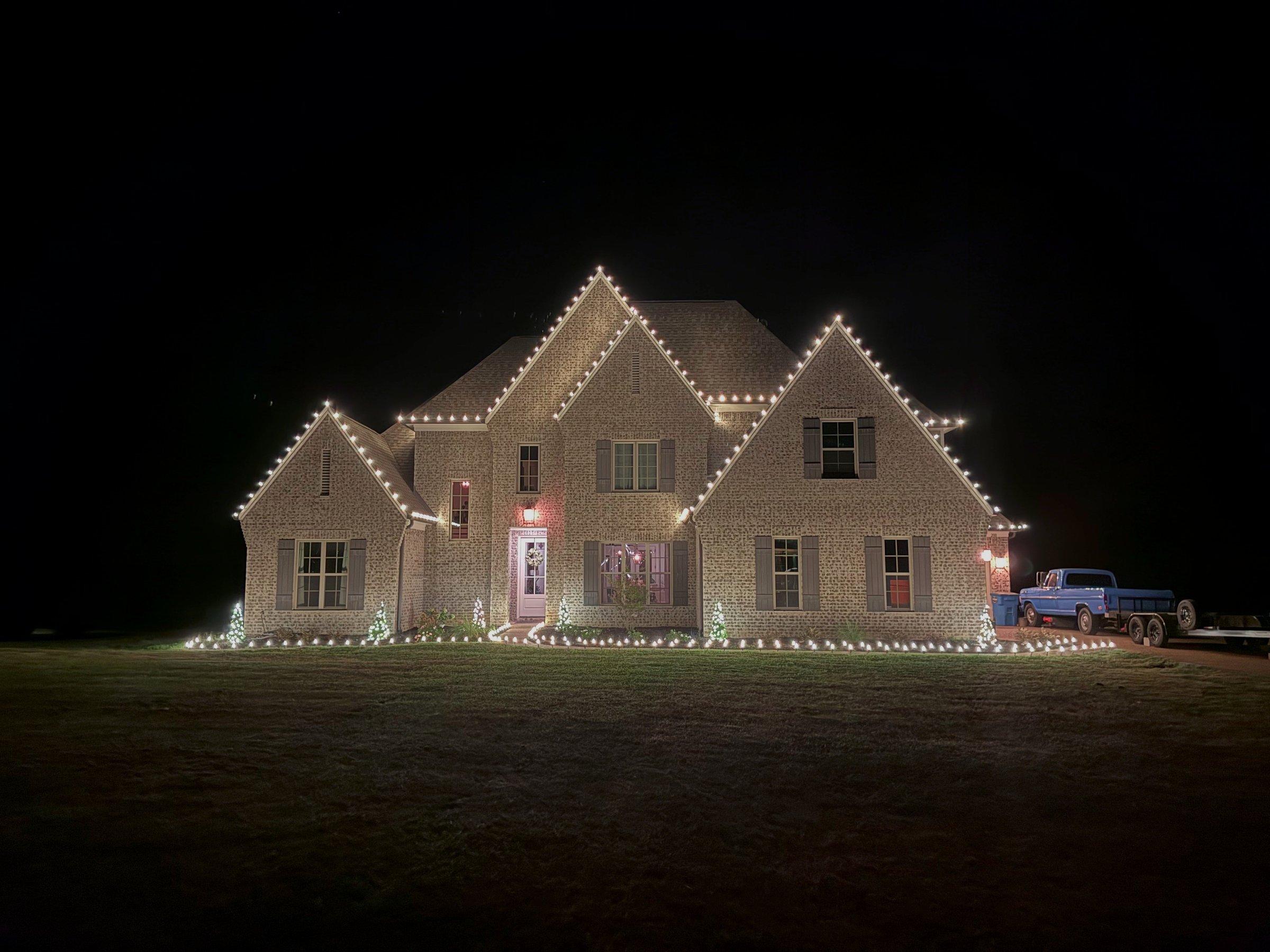 Two-story brick home at night fully outlined in warm white C9 bulbs along every roofline and gable, with a pickup truck and trailer parked in the driveway