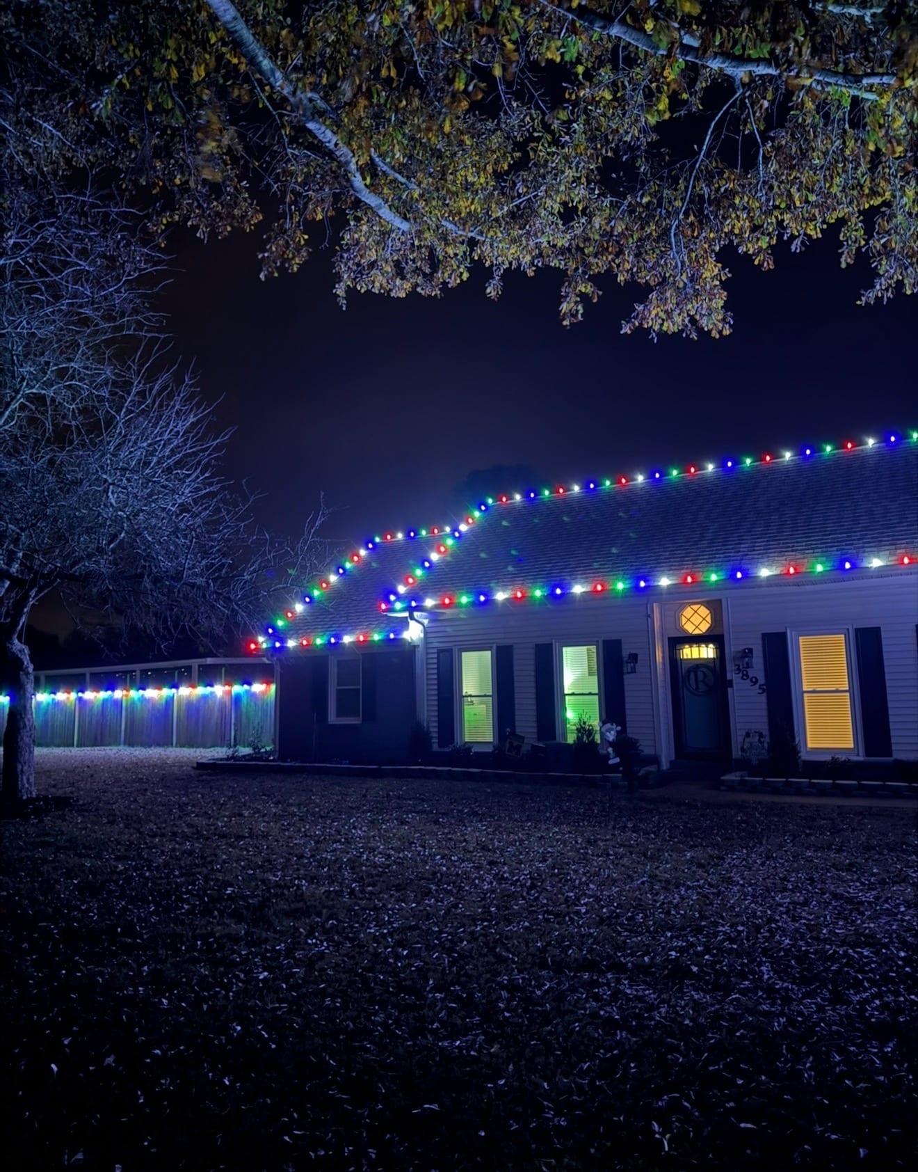 Home at night with red, green, blue, and white C9 bulbs along the roofline and a multicolor-lit fence line in the background