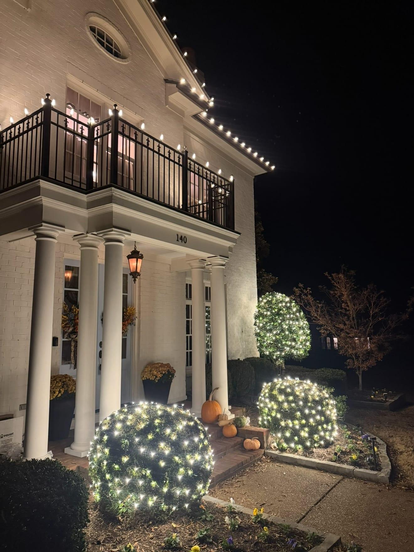 Boxwood shrubs net-lit into glowing spheres beside a white-brick home's columned entry, with a lit tree behind them