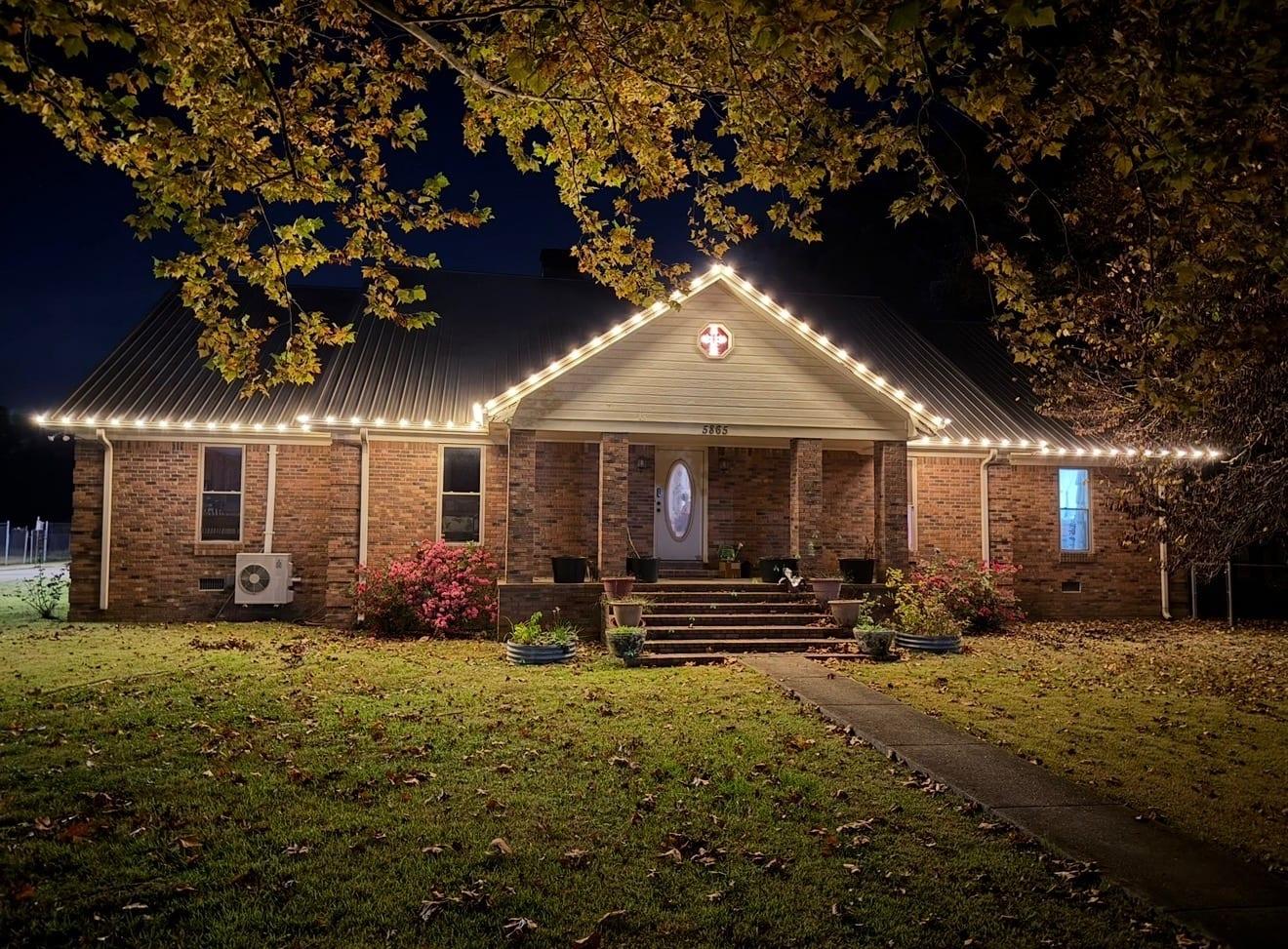 Brick ranch home at night with warm white C9 bulbs along the gabled roofline and porch, azalea bushes blooming below